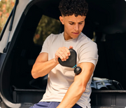 Man delivering percussive massage to his bicep with the Therabody Theragun Mini Plus at the back of his car