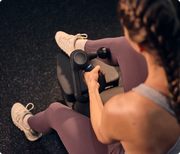 Woman delivering percussive massage to her calf on a gym bench using the Therabody Theragun Prime Generation 6