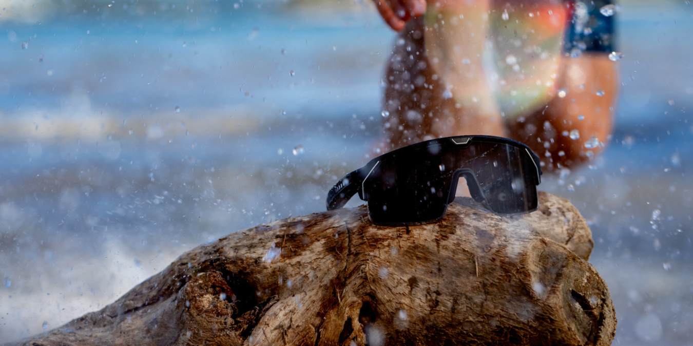 Chamelo Music Shield black frame smokey lenses on a rock with water splashing and a man in the background 